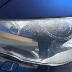 Głębokie rysy na reflektorze samochodu, matowa powierzchnia i liczne zadrapania.