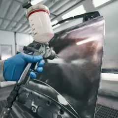 Ręka w niebieskiej rękawiczce trzyma pistolet lakierniczy, rozpylając substancję na metalowy element. To kluczowy etap prac, gdzie zastanawiamy się, czy na podkład epoksydowy można kłaść lakier.