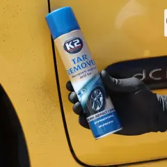 Rękawiczka z czarnym sprayem K2 Tar Remover na żółtym samochodzie. Skutecznie usuwa olej samochodowy i smołę.