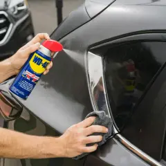 Usuwanie rys z lakieru wd40. Ręka trzyma puszkę WD-40 i spryskuje nią ciemny lakier samochodu, a druga ręka wyciera powierzchnię szmatką.