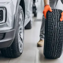 Mechanik w pomarańczowych rękawicach trzyma nowe opony asymetryczne obok szarego samochodu.