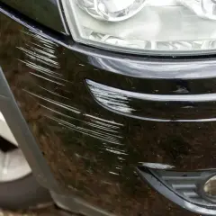 Głębokie otarcie zderzaka jak usunąć? Widoczne liczne rysy na czarnym lakierze, obok reflektora i fragmentu felgi.