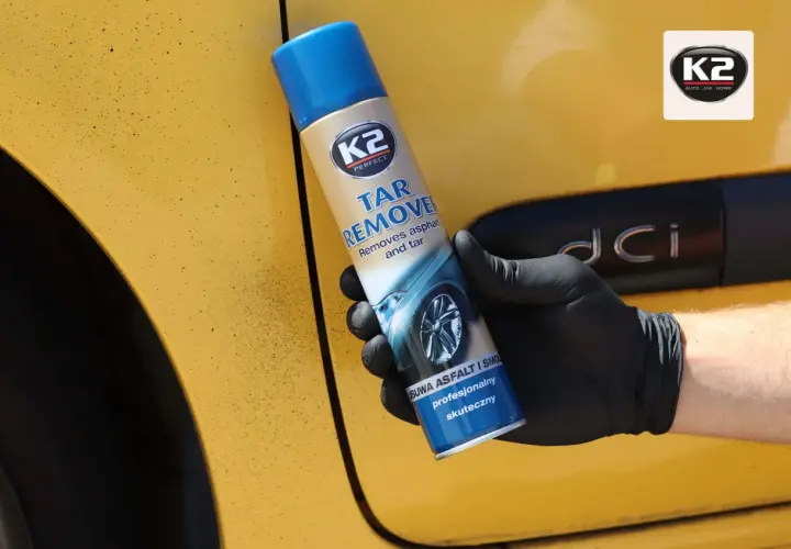 Rękawiczka z czarnym sprayem K2 Tar Remover na żółtym samochodzie. Skutecznie usuwa olej samochodowy i smołę.