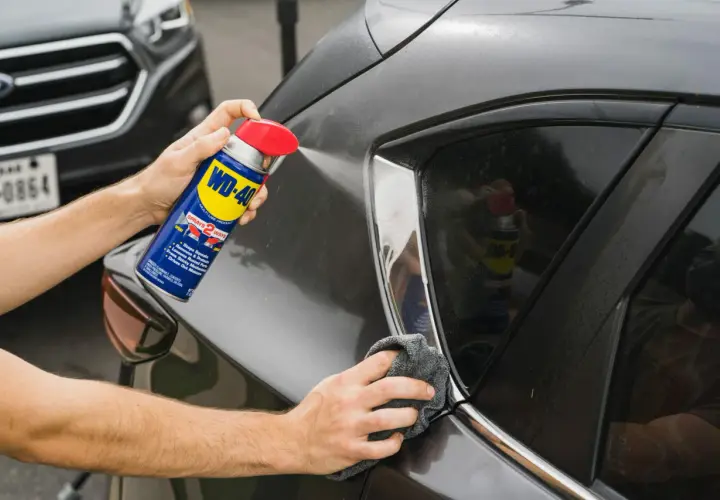 Usuwanie rys z lakieru wd40. Ręka trzyma puszkę WD-40 i spryskuje nią ciemny lakier samochodu, a druga ręka wyciera powierzchnię szmatką.