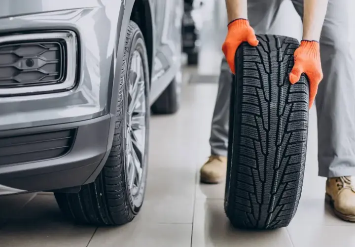 Mechanik w pomarańczowych rękawicach trzyma nowe opony asymetryczne obok szarego samochodu.