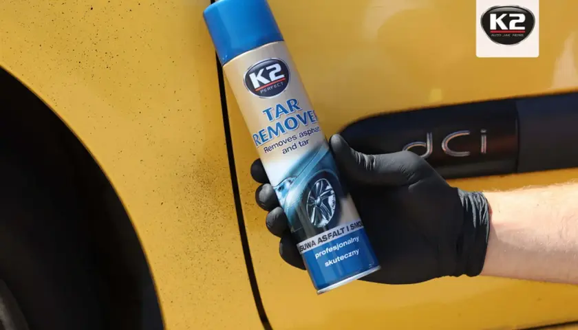 Rękawiczka z czarnym sprayem K2 Tar Remover na żółtym samochodzie. Skutecznie usuwa olej samochodowy i smołę.