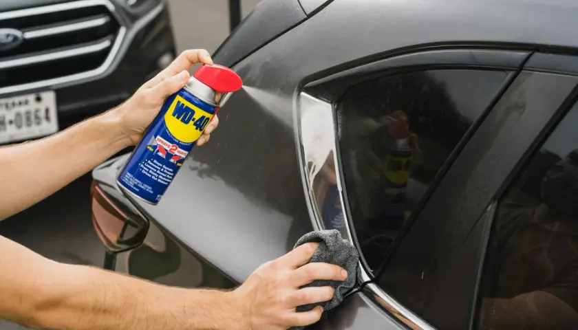 Usuwanie rys z lakieru wd40. Ręka trzyma puszkę WD-40 i spryskuje nią ciemny lakier samochodu, a druga ręka wyciera powierzchnię szmatką.