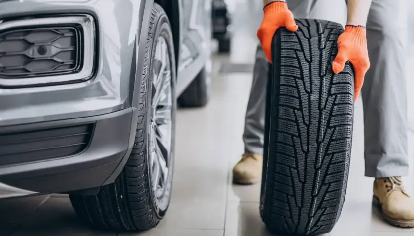 Mechanik w pomarańczowych rękawicach trzyma nowe opony asymetryczne obok szarego samochodu.