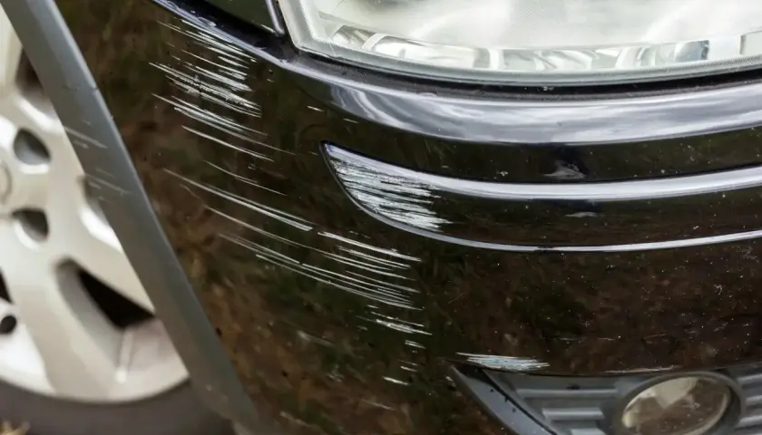 Głębokie otarcie zderzaka jak usunąć? Widoczne liczne rysy na czarnym lakierze, obok reflektora i fragmentu felgi.