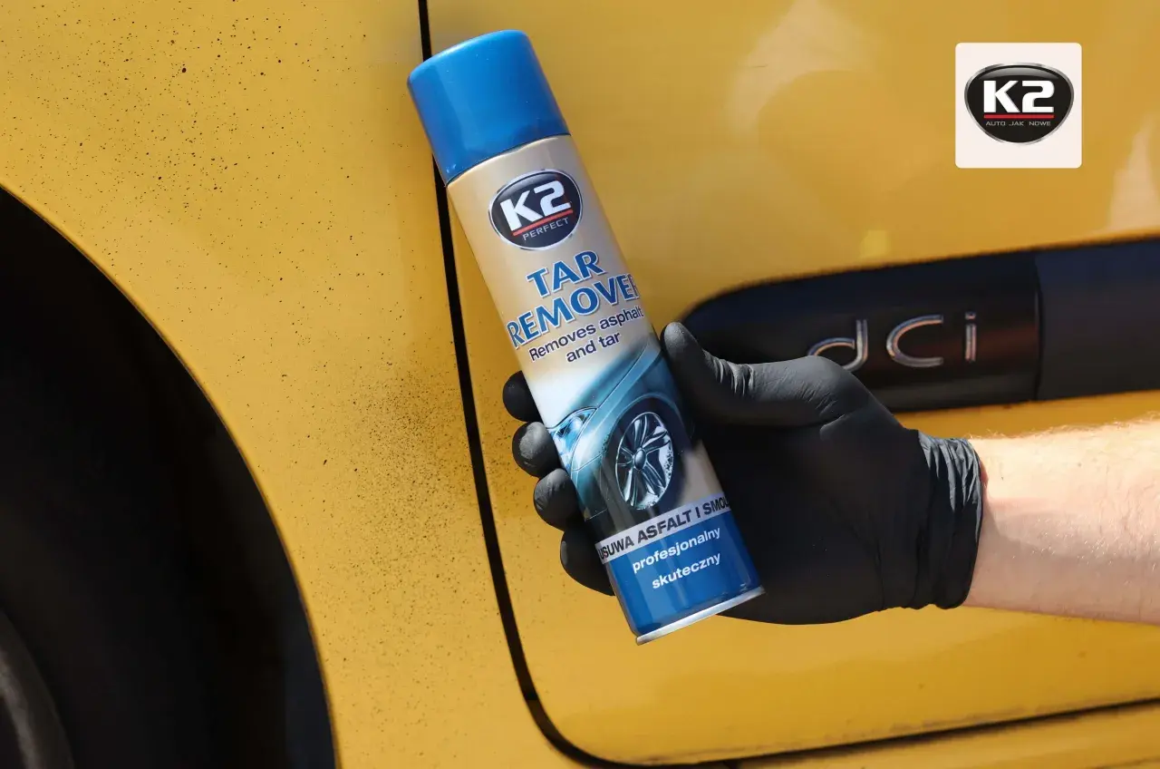 Rękawiczka z czarnym sprayem K2 Tar Remover na żółtym samochodzie. Skutecznie usuwa olej samochodowy i smołę.