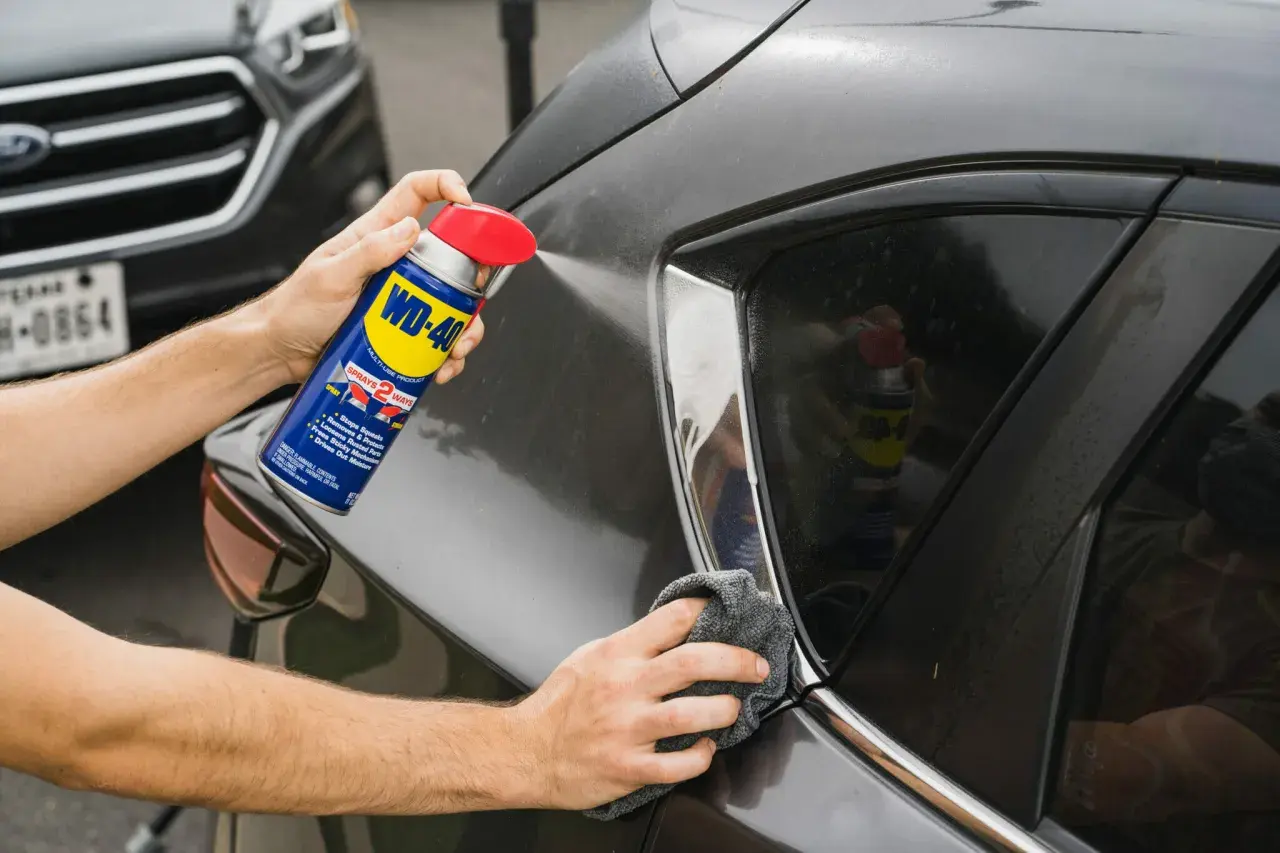 Usuwanie rys z lakieru wd40. Ręka trzyma puszkę WD-40 i spryskuje nią ciemny lakier samochodu, a druga ręka wyciera powierzchnię szmatką.