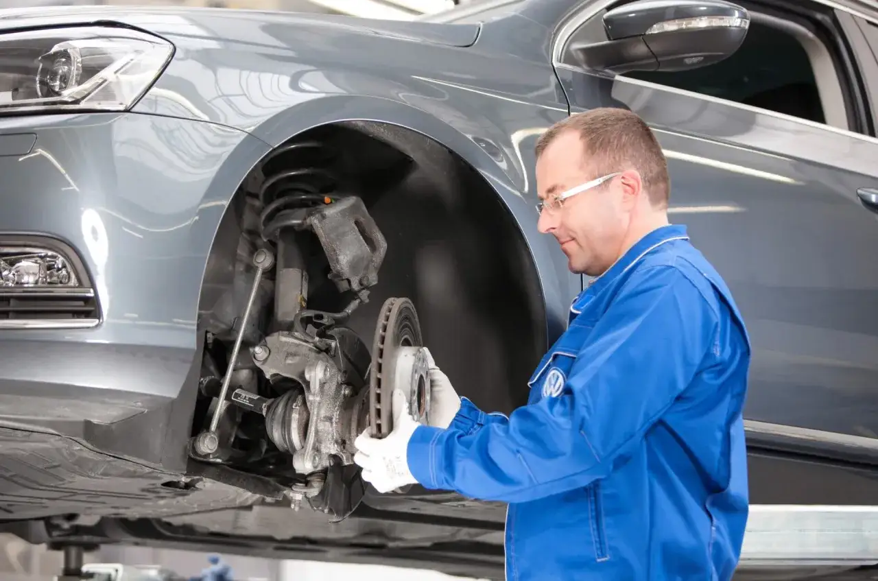 Mechanik w niebieskim kombinezonie wymienia tarczę hamulcową. To część kompleksowego serwisu samochodu, co obejmuje kontrolę i wymianę zużytych elementów układu hamulcowego.
