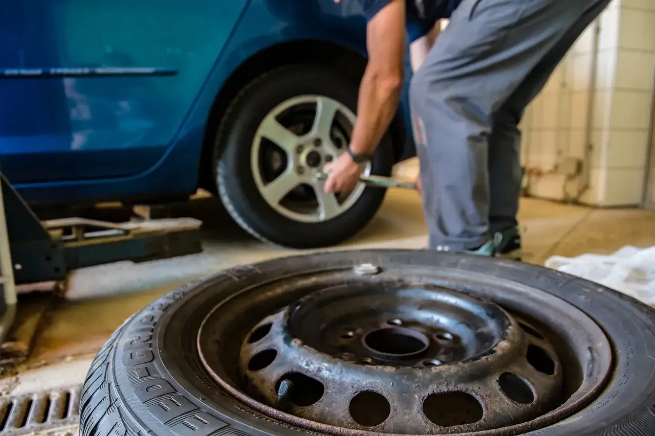 Mechanik dokonuje naprawy opon, odkręcając koło w niebieskim samochodzie. W tle leży zapasowa opona.